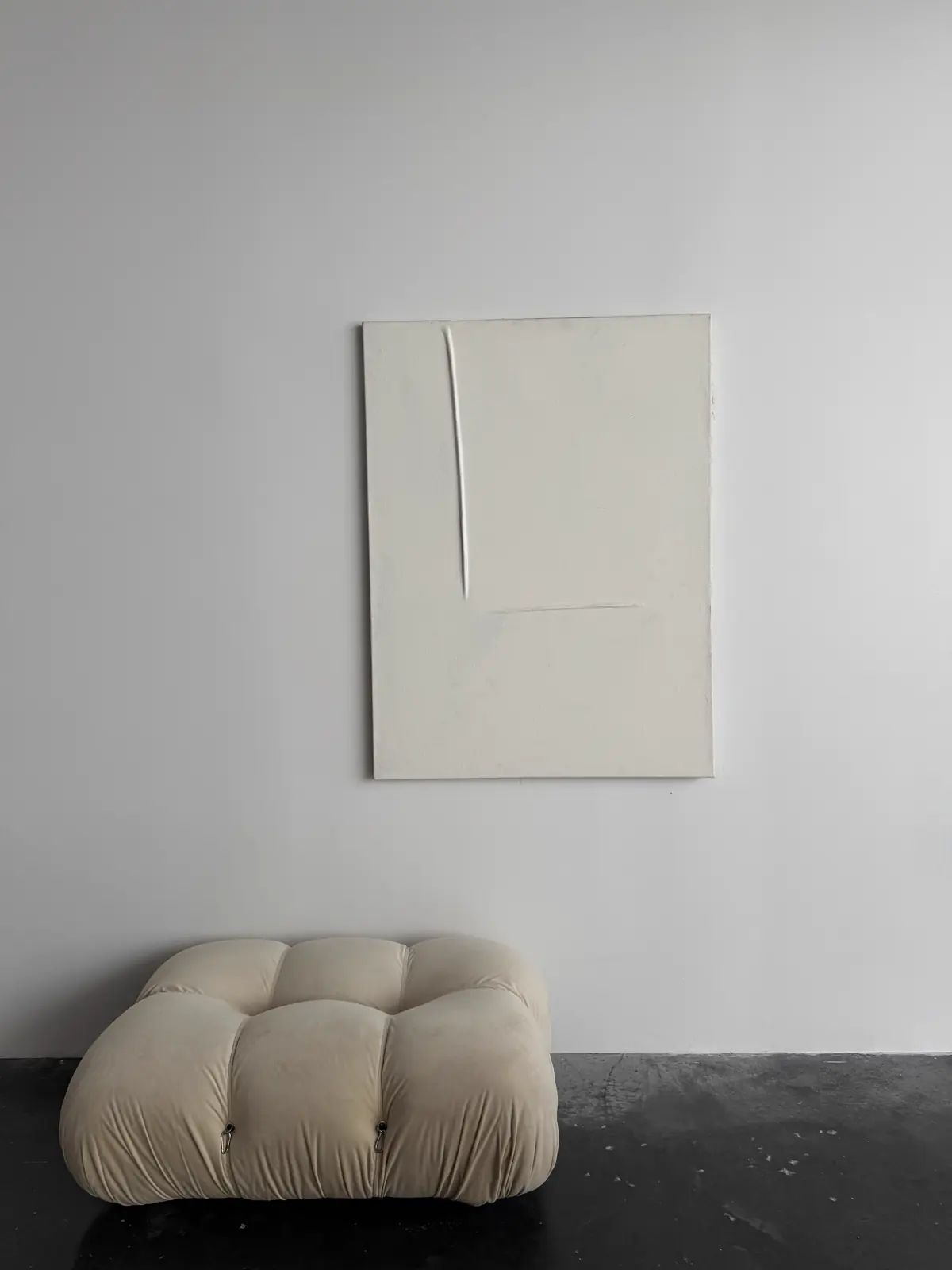 A large blank canvas hung above a cream pouf in an empty, evenly lit room