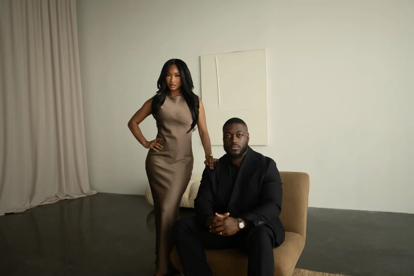 Aïcha standing in a satin dress beside Idriss, seated in black, in a cream studio
