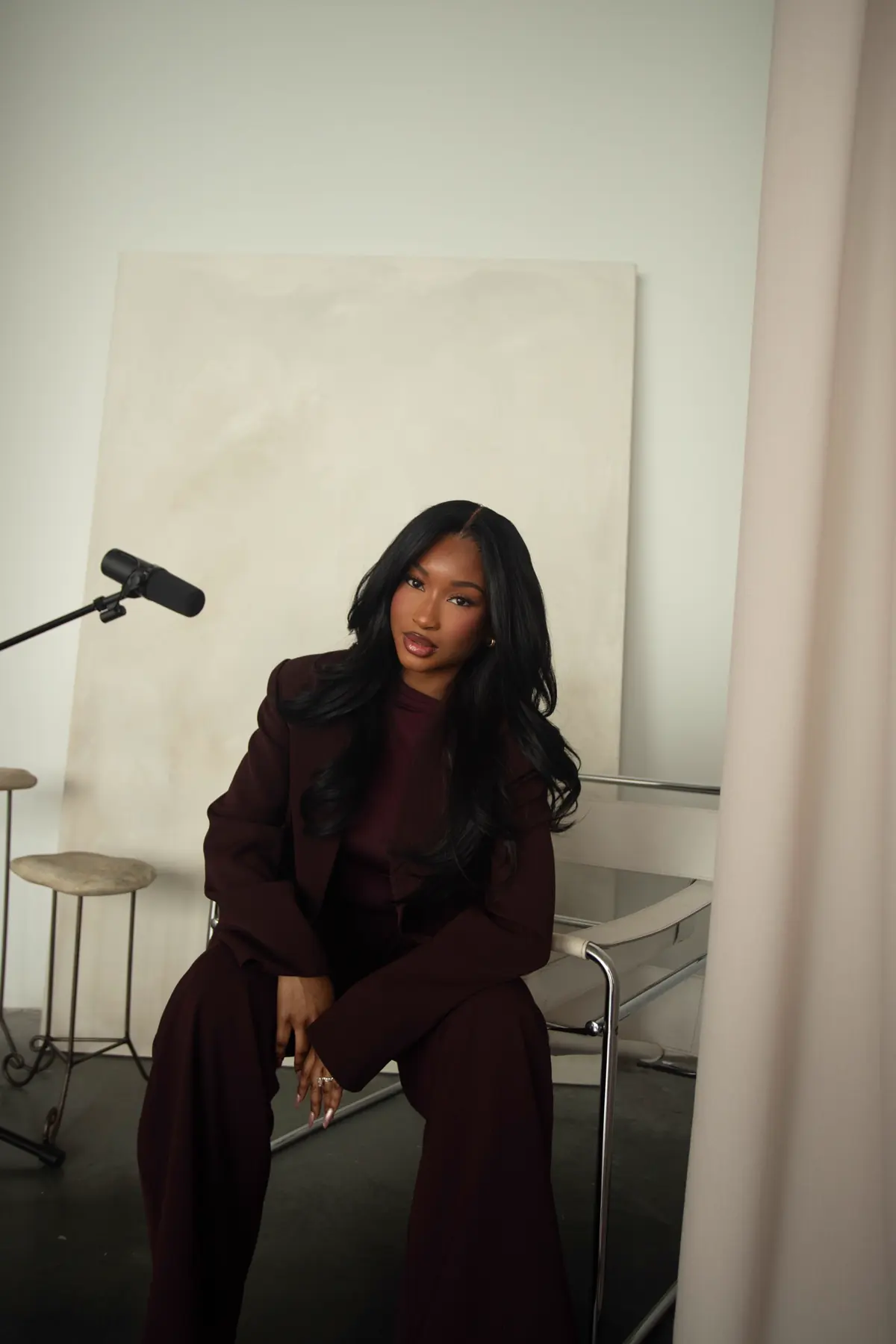 Aicha seated on set in a burgundy suit with a studio microphone in frame