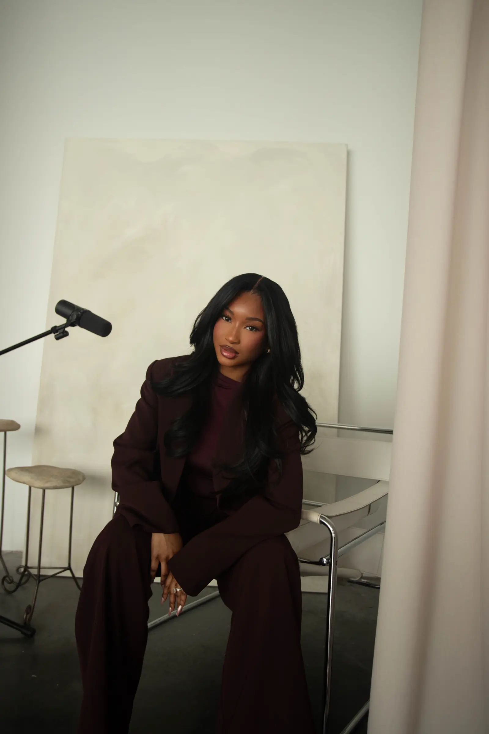 Aïcha seated on set in a burgundy suit, a studio microphone angled into the frame